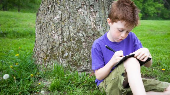 Little boy sitting in grass outside, writing in a notebook or journal.
