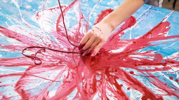 Close up of female painter bare hand creating a painting on canvas lying on the table in red, pink and blue color using rope covered in paint.