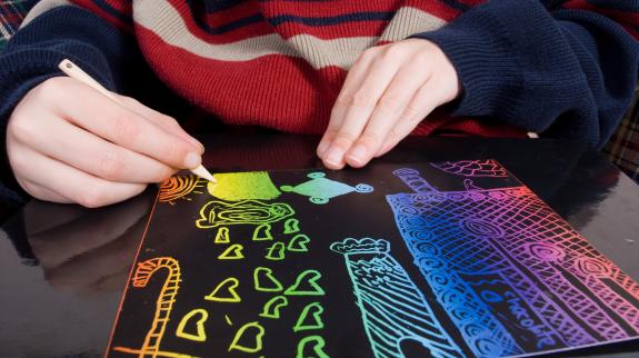 A young boy drawing a scene on scratch black paper.