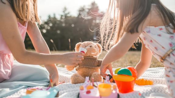 Two girls play with their teddy bears and have a picnic on summer day in park.
