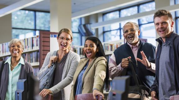 A multi-ethnic group of five adults standing in a library, looking up at the camera and smiling. 