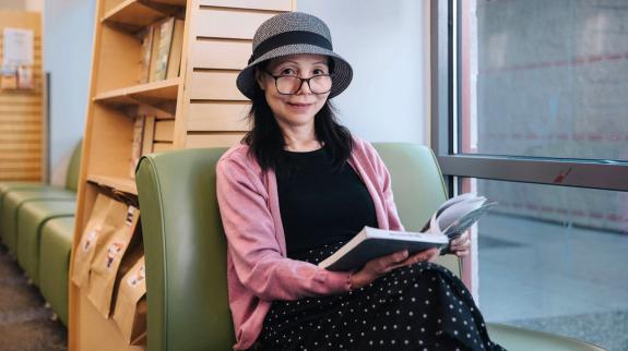 A woman sits reading a book at Dean Drysdale Library. A bookshelf with Grab & Go bags can be seen behind her.