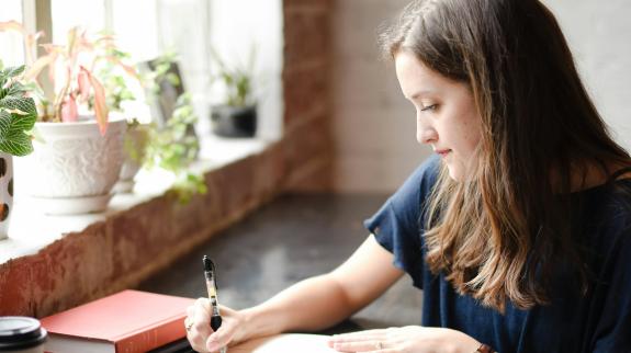 Woman writing in a notebook at a desk by the window.