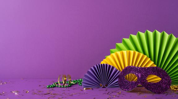 Side view colorful Mardi Gras accessories including a glittery purple mask, festive beads, and paper rosettes arranged on a bold purple background, capturing the celebratory essence of the festival.