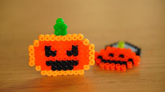 A close-up of two pumpkin shaped Perler charms lying side-by-side on a flat surface.