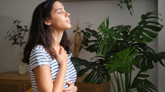 Young woman is taking a deep breath while standing in her living room, surrounded by plants.