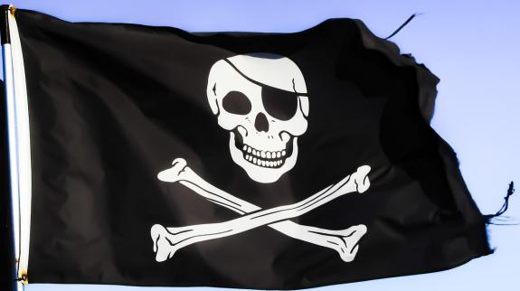 Black pirate flag has a white skull and crossbones with a black eyepatch and is rippling against a blue sky. The edges of the flag are slightly ripped.