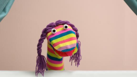 Rainbow sock puppet with purple braids and googly eyes performs against a peach background.