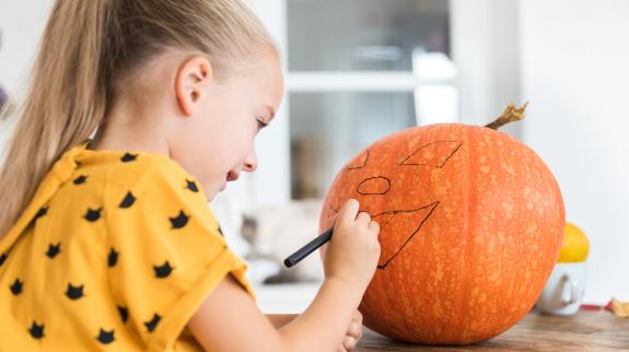 Girl sits at a table drawing a face on a large Halloween pumpkin. 