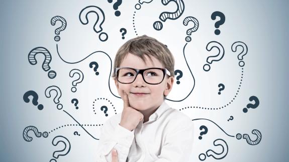 Smiling thinking little boy wearing glasses and white shirt standing near gray wall with many question marks drawn on it. 