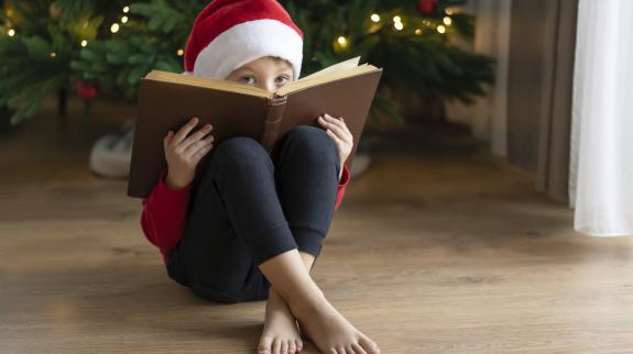 Child in Santa hat peeking over a book. A Christmas tree sits in the background.