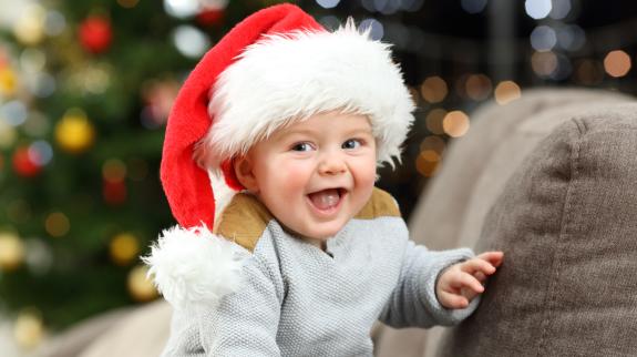 Baby with Santa hat sitting on couch smiling with Christmas tree in the background.