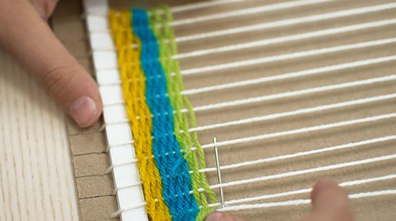 Pair of hands weave colourful yarn through the strings on a cardboard loom.