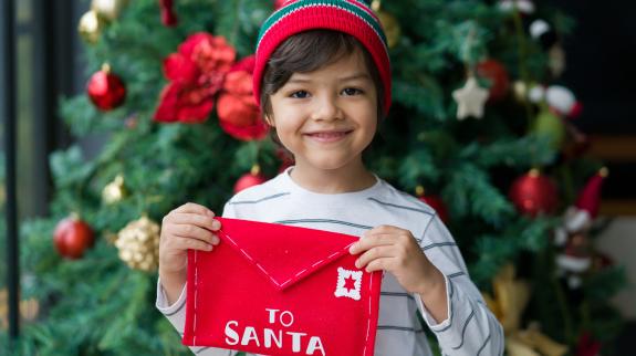 Boy holding a letter for Santa next to the Christmas Tree and looking at the camera smiling.