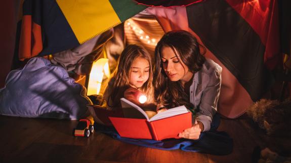 Mother reading a fairy tale book to her daughter under children's blanket fort at night.