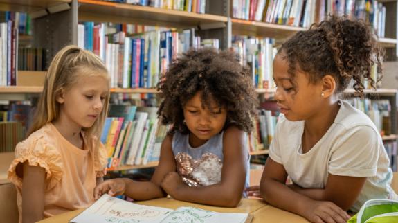 Three young girls read together at a library table.