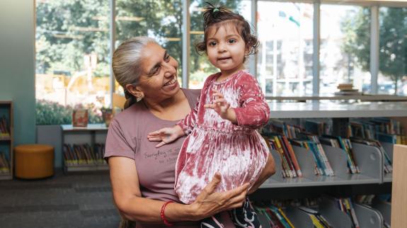 A South Asian woman poses for a photo. She holds her granddaughter, a toddler, in her arms. They have just arrived for Storytime at Terry Fox Library.