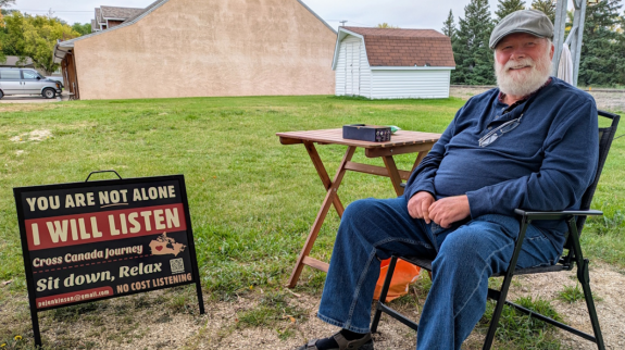 Bearded man sits in a chair with a table. Sign beside him says "I will listen."