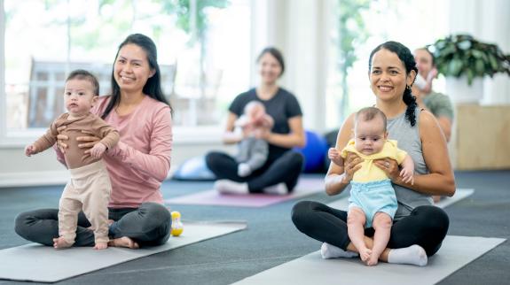 A small group of new parents sit on yoga mats in a studio, with their babies on their laps, as they participate in a parent & baby fitness class.