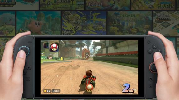 Hands holding a gaming console displaying a racing game on the screen. A character is driving on a dirt track, positioned in second place.