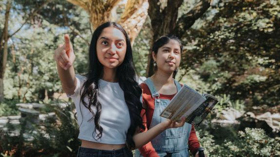 Two teenage girls explore the Abbotsford International Friendship Garden behind Clearbrook Library. They are holding a guide book and walking sticks from the FVRL Nature Pack, available from The Playground™ collection.