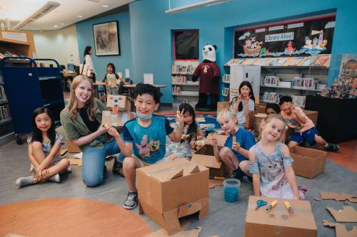 A large group of children and a female library staffer, cut up cardboard using Makedo tools and screws to create imaginative cardboard creations like a dragon head.