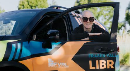 A woman in sunglasses leans out of a car, enjoying the breeze and sunshine with a cheerful expression.