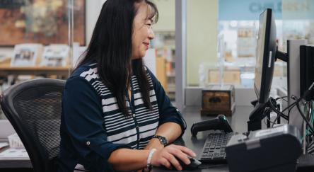 A librarian focused on her computer while sitting at a desk, surrounded by books and a warm, inviting library.