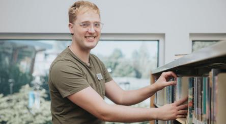 A young man in a green shirt smiles brightly while holding a book in his hands.