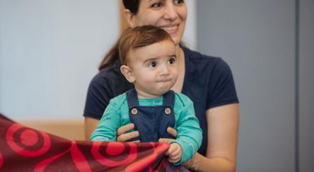 A woman holds a baby wearing a turquoise shirt and dark overalls. The baby looks ahead, holding a colorful fabric with a red and purple design.
