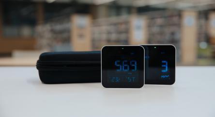 Two air quality monitors on a table in front of a library, displaying different air quality readings against a backdrop of bookshelves.