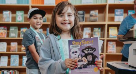 A young girl stands proudly in the library, clutching a book, eager to explore the world of stories.