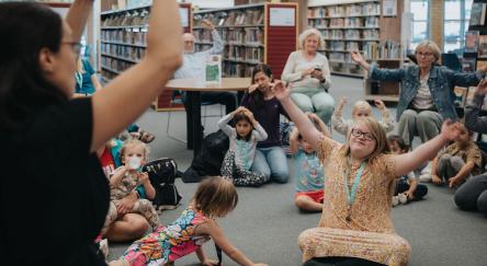 A lively scene in a library with children sitting and raising their hands, excited to participate in a group activity.