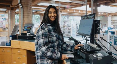A smiling woman stands at a library desk, surrounded by books, exuding warmth and friendliness.