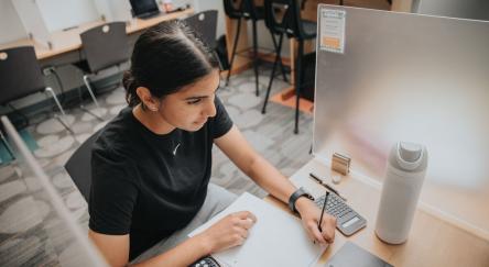 A woman sits at a desk, focused on her laptop, with a calculator beside her, ready to tackle her exam.