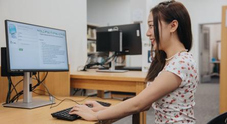  A woman sitting at a desk, focused on her computer screen, with a notepad and pen nearby.
