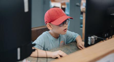  A young boy in a red hat and sunglasses sits at a computer, focused on the screen in front of him.