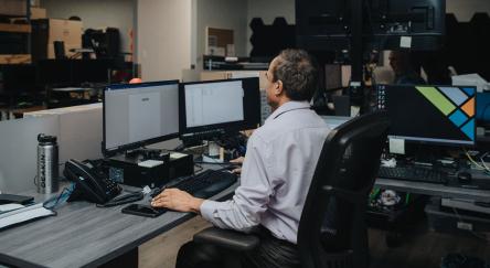  A man sits at a desk surrounded by multiple computer monitors, focused on his work.