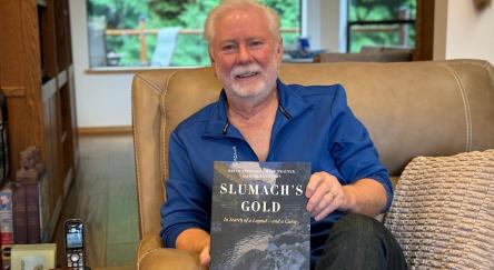 An older gentleman, author sits in a chair, showcasing the book, Slumach's Gold.