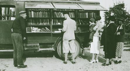 People of various ages stand around a vibrant bookmobile truck, discussing books and enjoying the community atmosphere.