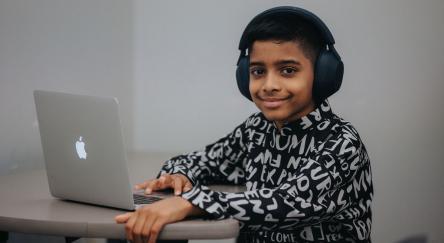 A boy works on a laptop computer in the library. He is wearing headphones.