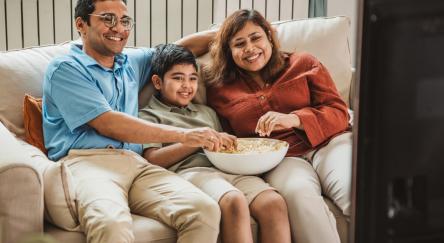A south asian family of three sits together on a couch, enjoying popcorn and a movie night in a cozy living room.