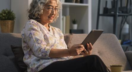 A senior woman sits on a couch, using a tablet, illustrating her comfort and engagement with digital technology.
