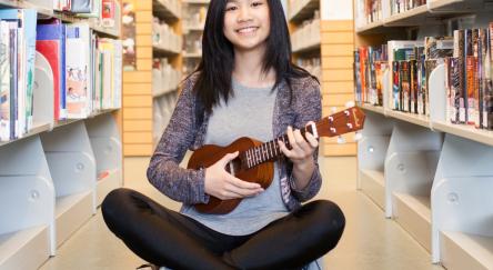 A young woman sits on the floor, strumming a ukulele with a joyful expression on her face.