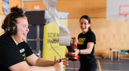 Two women joyfully interacting with a roll of toilet paper and leaf blower, showcasing creativity and laughter.
