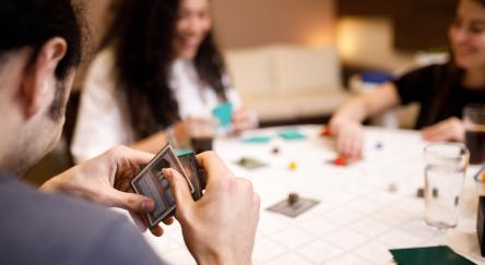 A man focused on his hand of cards, enjoying a table game with a smile on his face, surrounded by two friends.