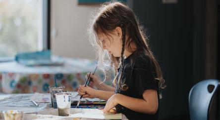 A young girl happily creating art at a table, immersed in her painting with a variety of colors and tools nearby.