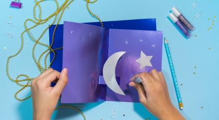 An overhead view of a pair of hands working on a purple and silver Ramadan craft. String, clips, and glitter glue sit on the table around the craft.