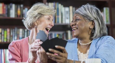 Two older women joyfully laughing together while each holding a hand of playing cards, sharing a moment of connection and friendship.
