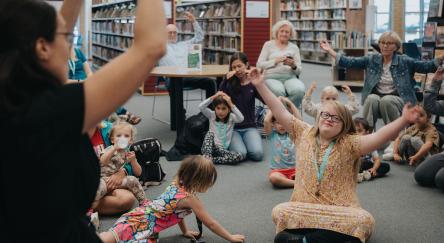 A lively scene in a library where children are seated, all raising their hands enthusiastically as they participate in Storytime.